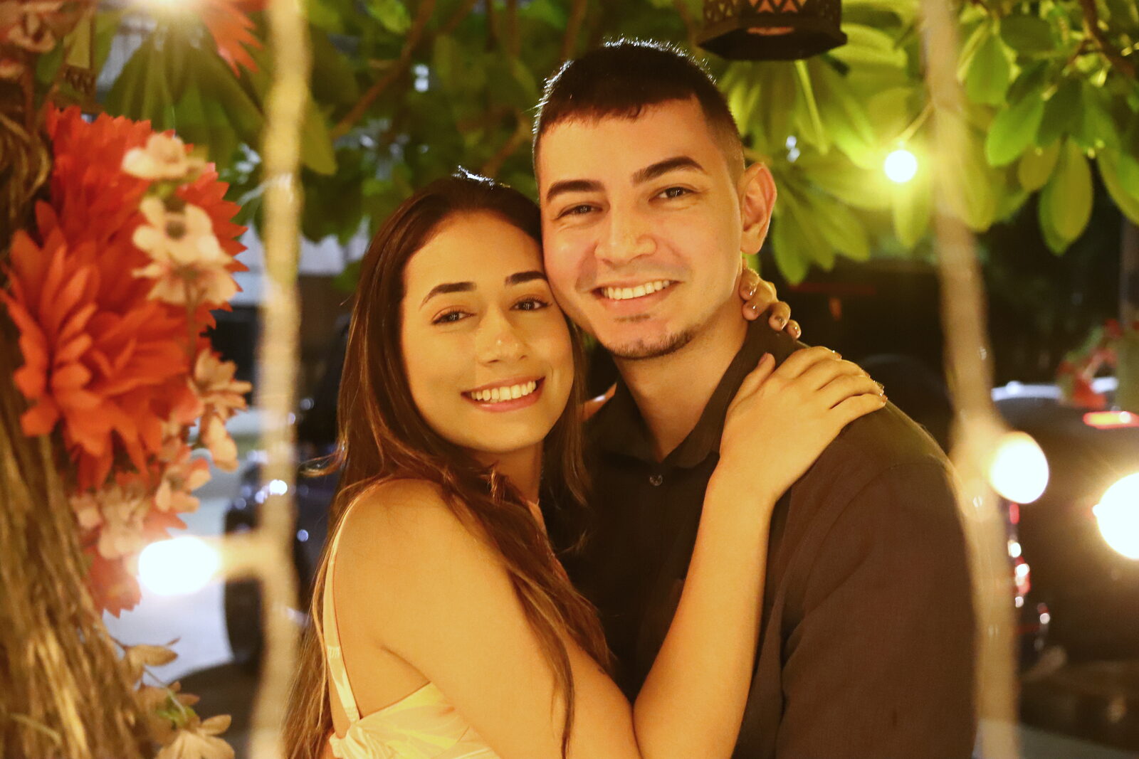 Danielle e Felipe abraçados à noite, sob luzes quentes e flores vermelhas